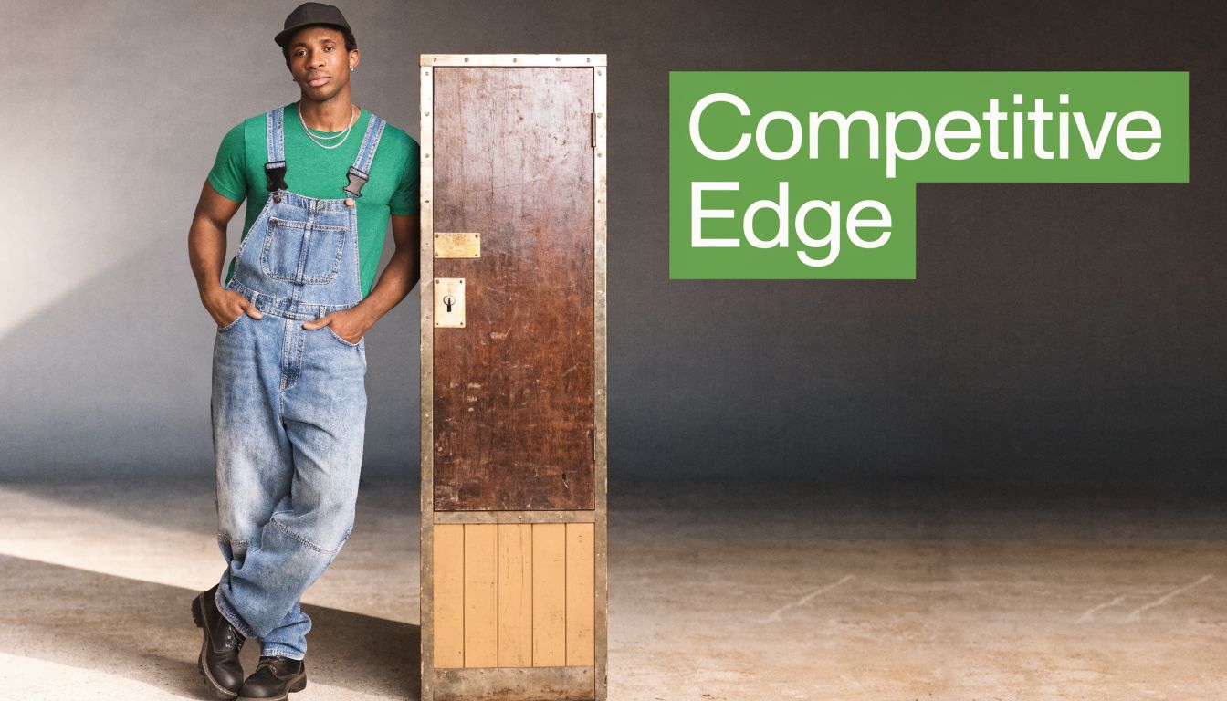 A young man wearing denim overalls and a green shirt leans against a tall wooden locker.